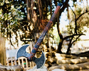 May include: A double-headed axe with a brown leather wrapped handle. The axe head is black and has a hammered finish. The axe is resting on a pile of logs in a wooded area.