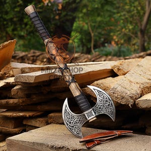 May include: A double-headed axe with a wooden handle wrapped in leather. The axe heads have intricate Celtic knotwork designs and are polished to a shiny finish. The axe is resting on a pile of wooden planks.