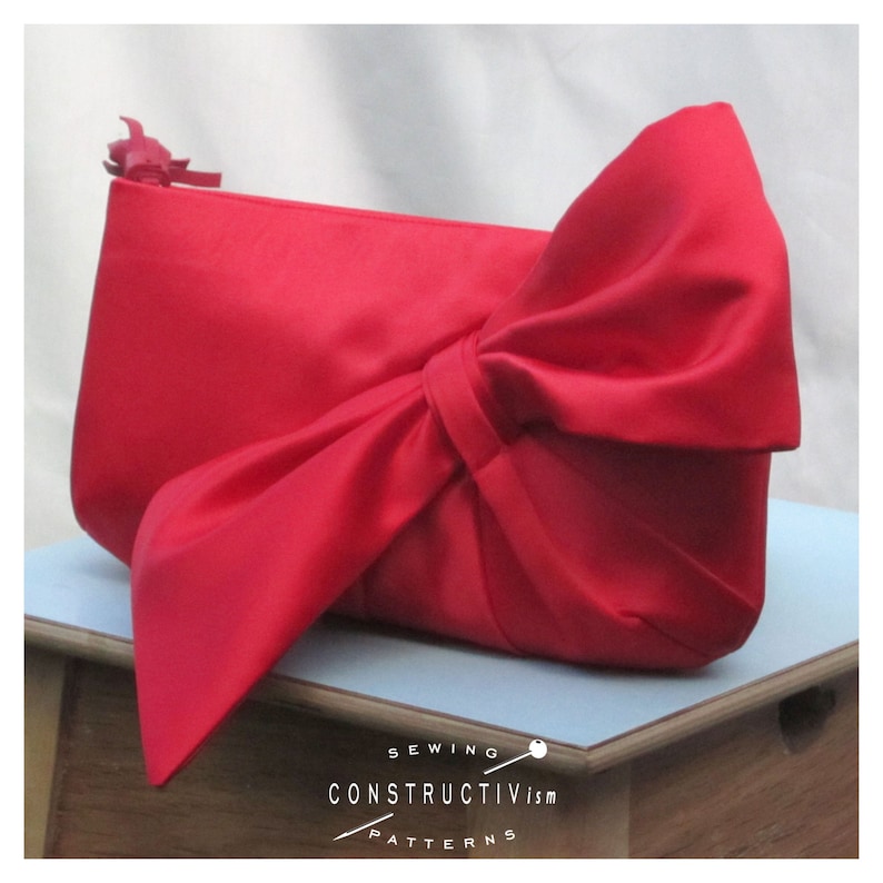 May include: A bright red satin clutch bag with a large bow on the front. The bag is sitting on a light blue surface.
