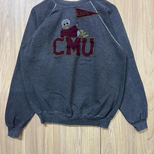 May include: Dark gray sweatshirt with a crew neck and long sleeves. Features a cartoon duck graphic with the text "CMU" in maroon and gold. A small flag with the word "CHIPS" is also present. The sweatshirt is hanging on a wooden hanger.