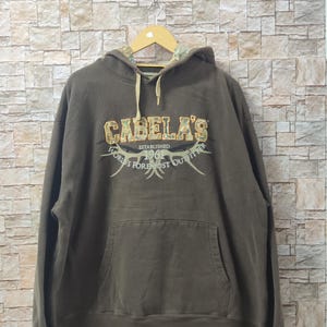 Puede incluir: Una sudadera con capucha marrón con un estampado de camuflaje en la capucha. La parte delantera de la sudadera tiene un gran logotipo bordado que dice "Cabela's Established 1961 World's Foremost Outfitter".