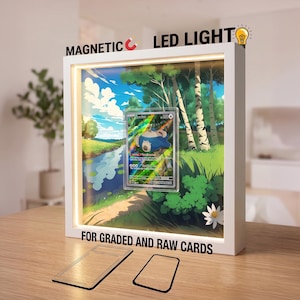 May include: A white square display frame with integrated LED lighting and a magnetic closure, featuring a Snorlax trading card inside. The frame displays a forest landscape background and includes text stating MAGNETIC LED LIGHT FOR GRADED AND RAW CARDS.