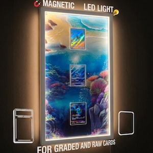 May include: A rectangular white magnetic LED light display frame featuring an underwater ocean scene with three trading cards mounted vertically. The text "MAGNETIC LED LIGHT" and "FOR GRADED AND RAW CARDS" is visible, along with icons of a magnet and a lightbulb.