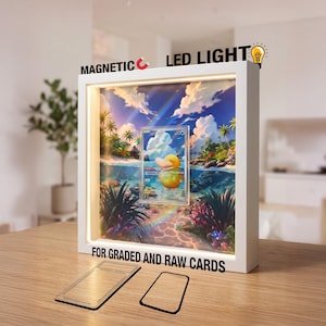 May include: A white square display frame with integrated LED lighting and a magnetic closure, designed for trading cards. The frame features a vibrant tropical beach scene background with a centered card, and includes two separate card inserts.
