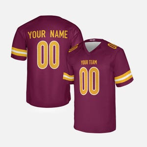 Puede incluir: Camiseta de fútbol burdeos con detalles dorados. La camiseta presenta el número "00" en dorado con contorno blanco, y los textos "YOUR NAME" y "YOUR TEAM" en dorado. Las mangas tienen rayas doradas y blancas.