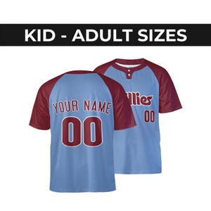 Può includere: Una maglia da baseball azzurra e bordeaux con maniche raglan e un'abbottonatura a due bottoni. La parte anteriore presenta la scritta Phillies e il numero 00. Il retro mostra la scritta YOUR NAME e il numero 00. Il testo recita KID - ADULT SIZES.