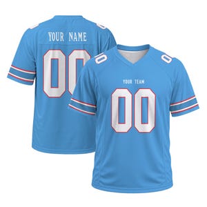 May include: A light blue sports jersey featuring white numbers 00 on the front and back, with white text reading YOUR TEAM on the chest and YOUR NAME on the back. The sleeves have red and white striped trim, and the V-neck collar is white.