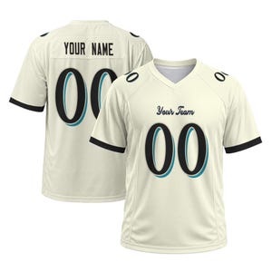 Peut inclure: Un maillot à manches courtes de couleur crème avec des accents noirs et bleu sarcelle. L'avant présente le texte "Your Team" et le numéro "00". Le dos affiche le texte "YOUR NAME" et le numéro "00". Les manches ont un bord noir.