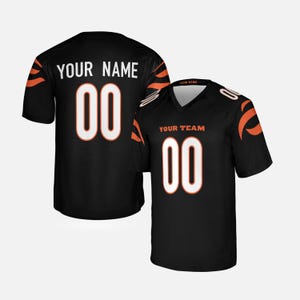 Könnte beinhalten: Ein schwarzes Sporttrikot mit orangefarbenen Tigerstreifenmustern auf den Schultern. Die Vorderseite zeigt den Text YOUR TEAM und die Nummer 00 in Weiß mit orangefarbenen Umrandungen. Die Rückseite zeigt den Text YOUR NAME und die Nummer 00 in Weiß.