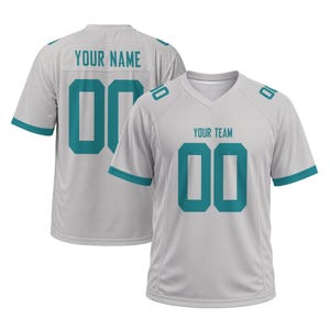 Puede incluir: Una camiseta deportiva de manga corta en color gris claro con ribetes en color verde azulado en las mangas y el cuello en V. El frente muestra el texto YOUR TEAM sobre el número 00 en verde azulado. La espalda lleva el texto YOUR NAME.