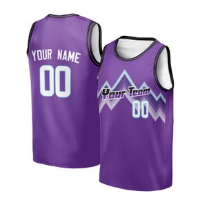 Peut inclure: Deux maillots de basket-ball violets. L'un porte "YOUR NAME" et le numéro "00" au dos. L'autre porte "Your Team" et le numéro "00" sur le devant, avec un motif de montagne. Les deux maillots ont des bordures noires et blanches.