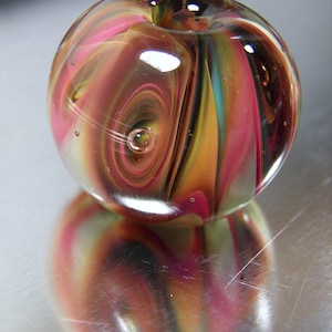 Peut inclure: Une bille de verre transparente avec des motifs tourbillonnants roses, oranges, jaunes et turquoise. La bille a une petite bulle d'air et se reflète sur une surface brillante. L'objet d'art en verre est rond et présente une petite indentation en haut.