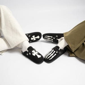 Może przedstawiać: Czarne zamszowe chodaki z białymi wzorami krzyży i kości szkieletu. Chodaki mają regulowane paski i są noszone z białymi skarpetami i spodniami casual. Obuwie jest prezentowane na białym tle.