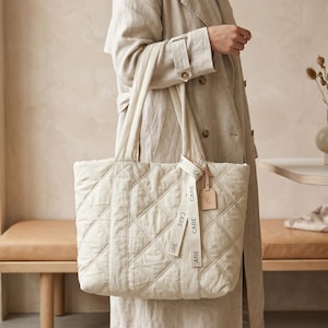 Peut inclure: Sac fourre-tout matelassé crème avec surpiqûres en losange. Le sac a de longues anses et une étiquette avec la lettre "K". Un ruban avec le mot "CARE" est attaché au sac. Le sac est tenu par une personne portant un trench-coat beige.