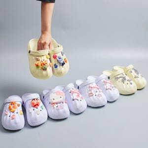 May include: A collection of light-coloured clogs featuring various decorative charms. The footwear includes white and cream-coloured slip-on shoes with textured surfaces, adorned with colourful character figures, bows, and small icons attached to the ventilation holes.