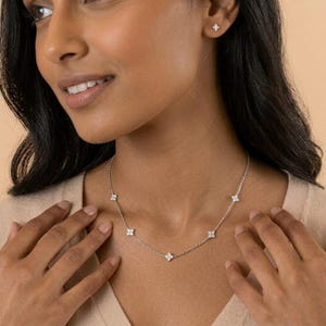 May include: A silver necklace and matching earrings featuring a floral design with clear gemstones. The necklace has five floral pendants, and the earrings have one floral stud each. The jewelry is worn by a person wearing a beige top.