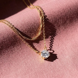 May include: A gold necklace featuring a large, clear, round gemstone pendant. The necklace has a fine chain and is set against a pink fabric background. The pendant is the central feature of the jewellery.