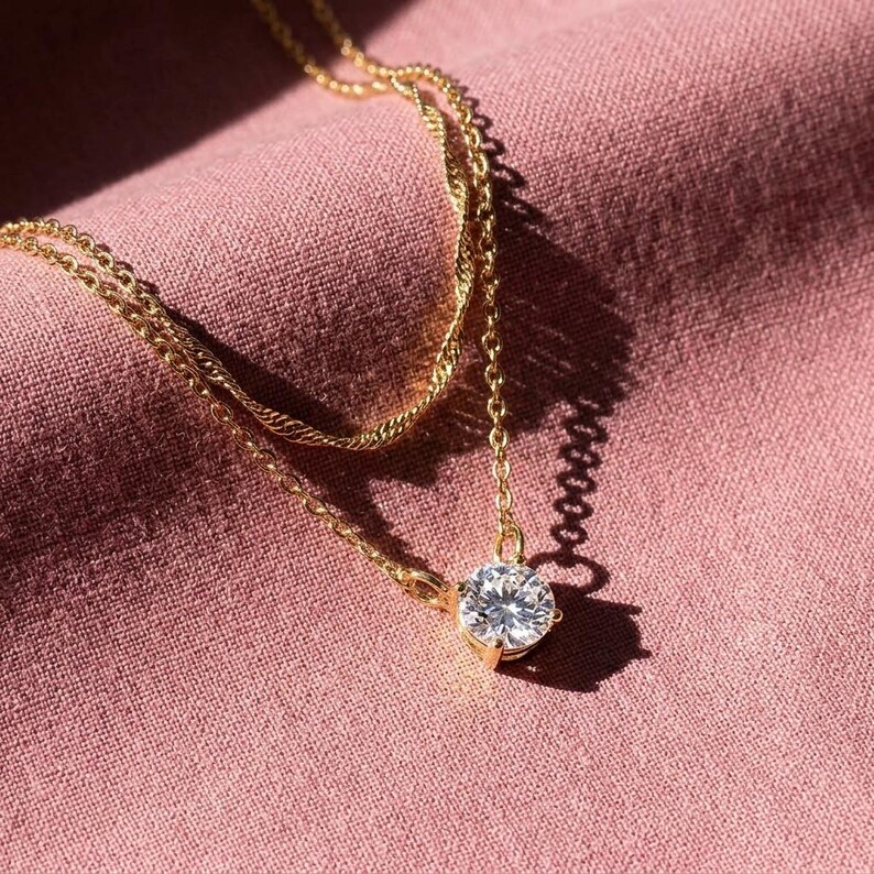 May include: A gold necklace featuring a large, clear, round gemstone pendant. The necklace has a fine chain and is set against a pink fabric background. The pendant is the central feature of the jewellery.