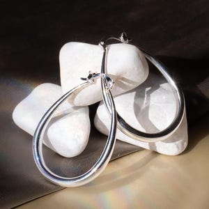 May include: A pair of silver teardrop hoop earrings. The earrings are displayed on white stones against a dark background. The hoops have a smooth, polished finish and a classic design. The earrings are a stylish accessory.