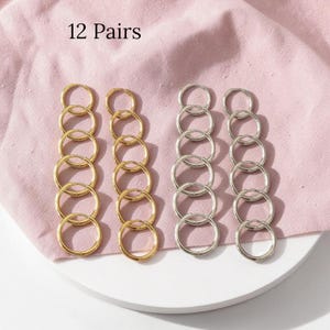 May include: An assortment of 12 pairs of hoop earrings in gold and silver tones. The earrings are arranged in rows on a pink fabric. The text "12 Pairs" is at the top of the image.