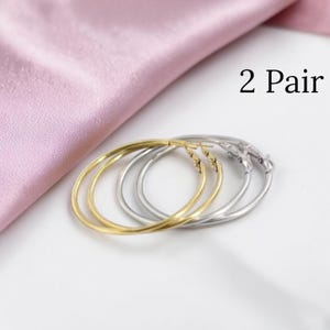 Op de afbeelding: Twee paar oorringen, één goudkleurig en één zilverkleurig, liggen op een witte ondergrond naast een roze satijnen stof. De tekst "2 Pair" is zichtbaar. De oorbellen zijn eenvoudig en elegant.