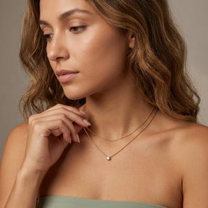 May include: A gold necklace with two strands. The top strand is a delicate chain, and the bottom strand features a small, round, clear gemstone pendant. The woman in the image is wearing a green top.