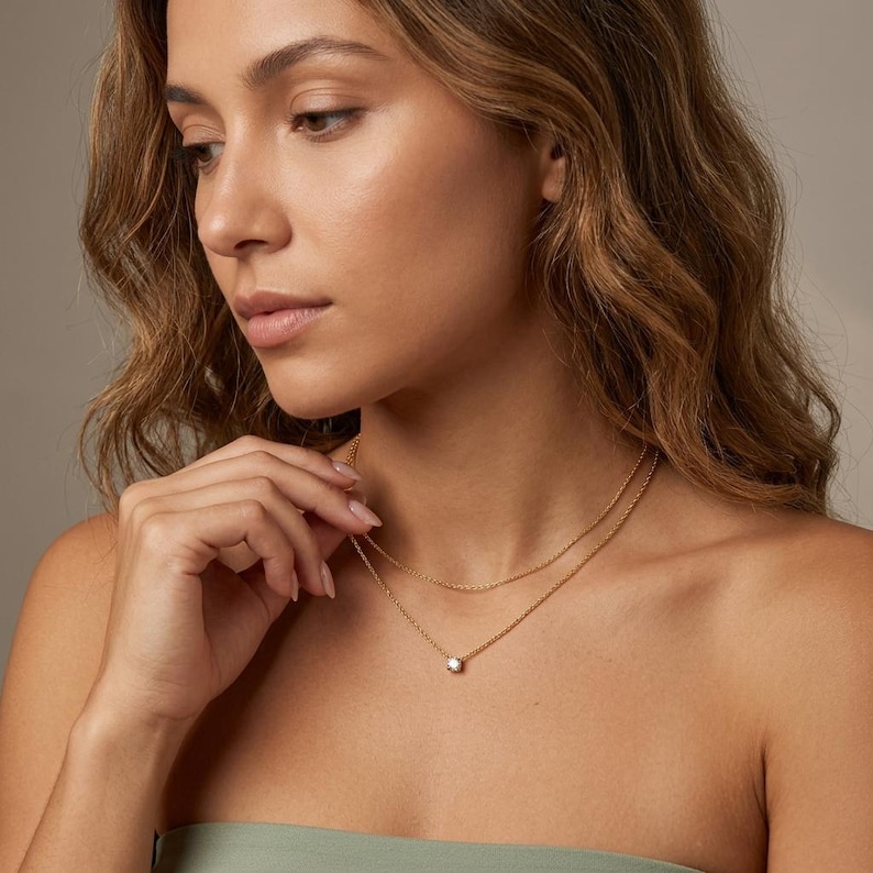 May include: A gold necklace with two strands. The top strand is a delicate chain, and the bottom strand features a small, round, clear gemstone pendant. The woman in the image is wearing a green top.