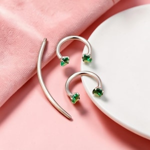 May include: Silver-toned horseshoe earrings with emerald green gemstones. A silver bar earring is also present. The jewelry is displayed on a pink surface with a white plate in the background. The earrings have a modern, minimalist design.