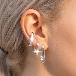 May include: Three silver-toned hoop earrings in a rectangular, elongated shape. The earrings are arranged in a vertical row along the earlobe and cartilage, featuring a polished, reflective metallic finish with a hinged closure mechanism.