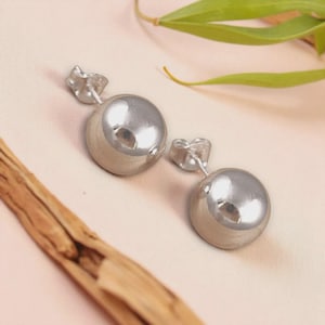 May include: A pair of shiny, silver-colored ball stud earrings. The earrings have a smooth, reflective surface and are set against a soft, light pink background with a wooden accent and green leaves.