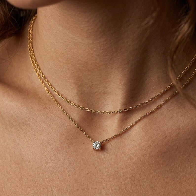 May include: A gold necklace with two layers. The top layer is a rope chain, and the bottom layer has a single, round, clear gemstone pendant. The necklace is shown on a person's neck.