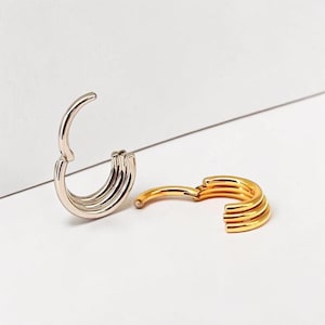 Può includere: Due orecchini a cerchio, uno argentato e uno dorato, sono mostrati su sfondo bianco. Ogni orecchino ha un design a cerniera e un aspetto multistrato e scanalato. Gli orecchini sono aperti, mostrando la loro struttura interna.