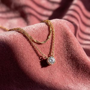 May include: A gold necklace featuring a large, round, clear gemstone pendant. The necklace has a delicate chain design and is displayed on a pink velvet surface. The necklace is a piece of jewellery.