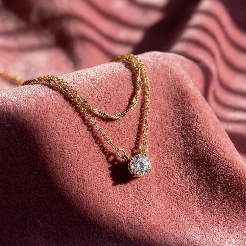 May include: A gold necklace featuring a large, round, clear gemstone pendant. The necklace has a delicate chain design and is displayed on a pink velvet surface. The necklace is a piece of jewellery.