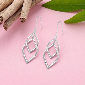 May include: A pair of silver-colored drop earrings. Each earring features a double, open, twisted design. The earrings hang from a simple hook. The background is a soft pink.