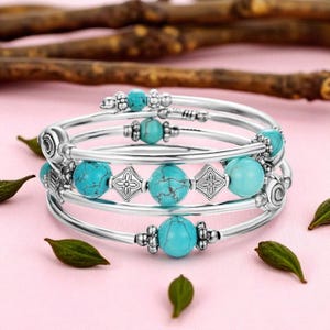 May include: Silver-toned bangle bracelets with turquoise-colored beads and decorative silver accents. The bracelets are stacked, showcasing the design and details of the jewelry. The background is a soft pink with scattered green leaves and wooden twigs.