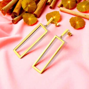 Könnte beinhalten: Ein Paar goldfarbene, rechteckige Ohrhänger. Die Ohrringe werden auf einer rosa Oberfläche präsentiert, mit getrockneten Stielen und gelb-orangefarbenen, blütenartigen Objekten im Hintergrund. Schlichtes, modernes Design.