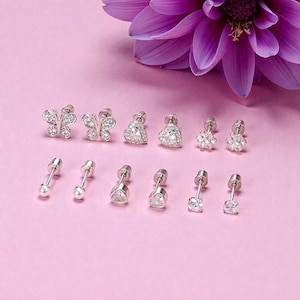May include: A collection of silver-toned stud earrings with various designs, including butterflies, hearts, and flowers, each adorned with clear gemstones. Some earrings feature a single pearl. The earrings are arranged on a pink surface, with a purple flower in the background.