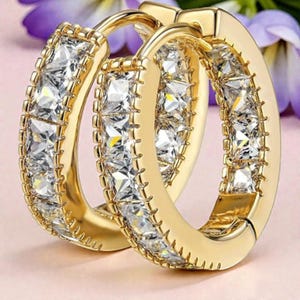 May include: A pair of gold-colored hoop earrings. Each earring is encrusted with square-cut, clear gemstones. The earrings are set against a soft pink background with a hint of purple flowers, highlighting the jewelry's sparkle.