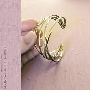 May include: A gold-toned cuff bracelet featuring a multi-strand design with four thin, polished metal bands that overlap and intersect to create a layered, open-ended structure. The metallic surface reflects light, highlighting the smooth, curved lines.