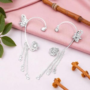May include: A pair of silver ear cuffs with butterfly and floral designs. Each cuff features a butterfly, a flower, and dangling chains with small, round embellishments. The jewelry is displayed on a pink surface.