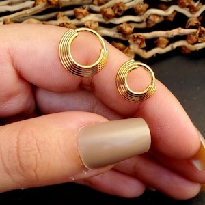 May include: Two gold-colored hoop earrings with a layered design. The earrings are made of multiple thin hoops stacked on top of each other.