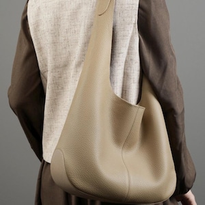 May include: A beige leather shoulder bag with a curved design. The bag has a single strap and a spacious interior. The bag is worn over the shoulder. The bag is made of a soft, textured leather.