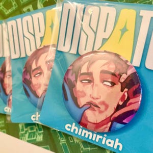 May include: A collection of round buttons featuring a stylized portrait of a person with a blue and purple color scheme. The buttons are packaged on blue cards with the word "DISPATCH" and the name "chimiriah" printed on them.