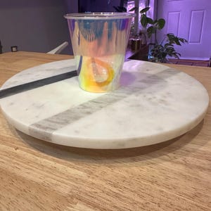May include: A clear iridescent plastic cup sits on a white marble round serving tray. The tray is on a wooden surface.