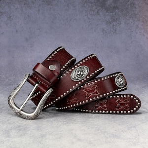Può includere: Una cintura in pelle marrone scuro con borchie metalliche argentate lungo i bordi, una fibbia incisa decorata e conchos ovali con motivi a stella. Il cinturino presenta una serie di intagli a forma di X e dettagli di cucitura.