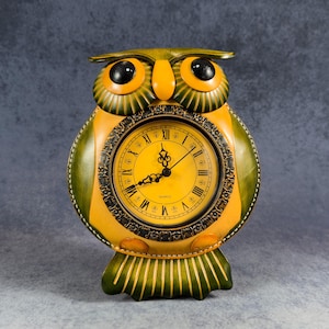 May include: A yellow and green owl-shaped clock with black Roman numeral markings and a quartz movement. The clock has a black face with black hands.
