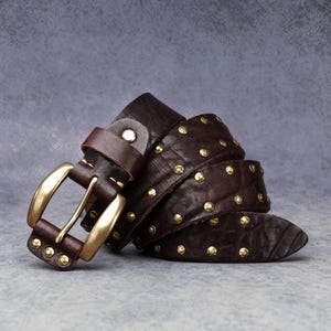 May include: A dark brown leather belt featuring a polished brass buckle and evenly spaced gold-toned metal studs along the strap. The belt is coiled, showcasing the textured surface of the leather and the metallic hardware details throughout.