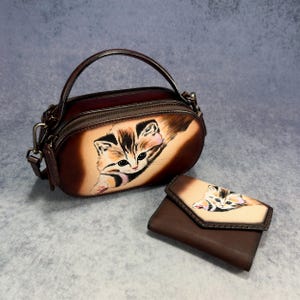 May include: A brown leather handbag and matching wallet. The handbag is oval-shaped with a top handle and shoulder strap. Both items display a painted kitten with brown, black, and tan markings.