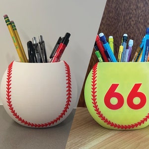 Puede incluir: Dos porta lápices con temática de béisbol. El de la izquierda es blanco con costuras rojas y está lleno de lápices y bolígrafos. El de la derecha es amarillo con costuras rojas y el número 66 en la parte delantera. También está lleno de lápices y bolígrafos.
